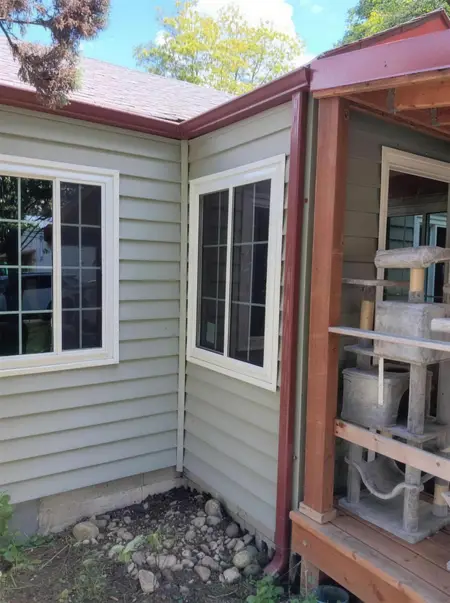 Corner of a beige house with two new windows and a red gutter line beside a covered porch