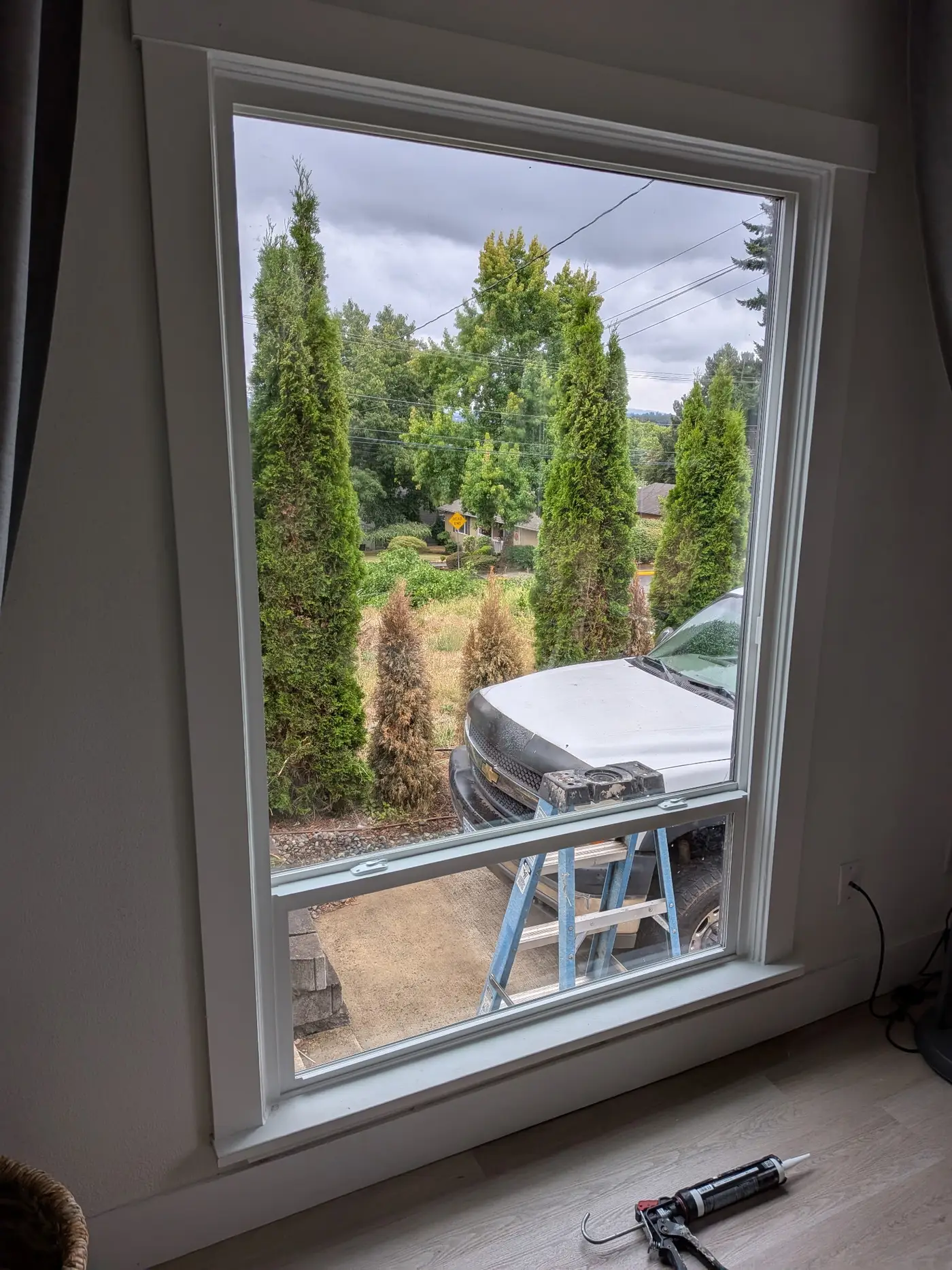 Caulk gun resting on the sill of a newly set window, seen from inside looking out at the job truck and ladder