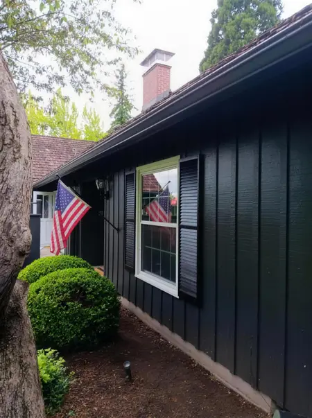 Window with black shutters on a dark green house beside American flags and a boxwood shrub
